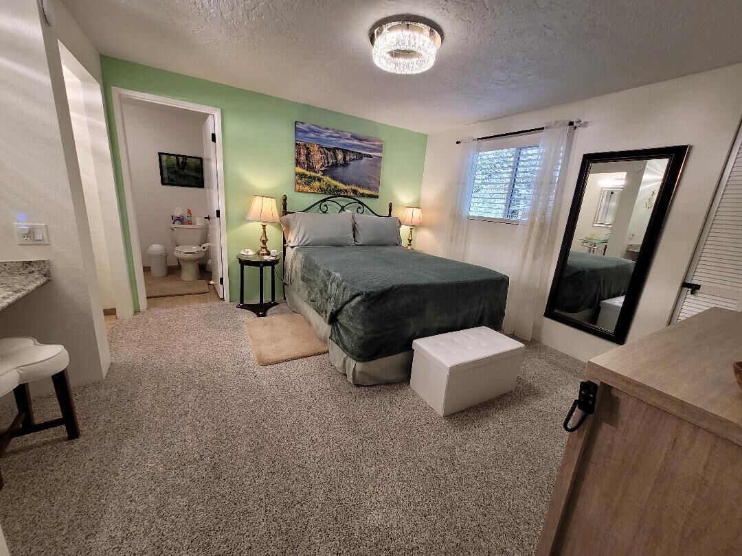 Une chambre à coucher avec un lit, une table de chevet et un miroir. On peut apercevoir la salle de bain à travers une porte ouverte.