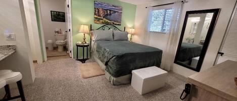 Une chambre à coucher avec un lit, une table de chevet et un miroir. On peut apercevoir la salle de bain à travers une porte ouverte.