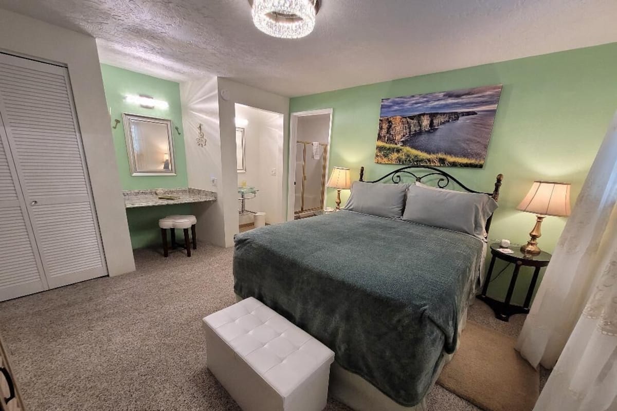 A bedroom with a bed, a nightstand, a lamp, a bench, a mirror, and a painting on the wall.
