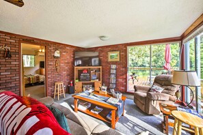 Ein gemütliches Wohnzimmer mit Backsteinwand, Fernseher, Bücherregal und großem Fenster mit Blick auf Grünflächen.