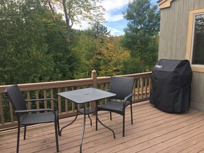 A wooden deck with two chairs and a table, a grill, and a view of trees and hills.