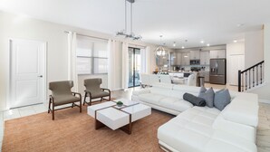 A modern living room with a white sofa, two armchairs, and a coffee table. A kitchen is visible in the background with white cabinets and stainless steel appliances. - 6BR Luxury Villa: Pool, Spa, Game Room & Disney Magic 2957 FS (Kissimmee)