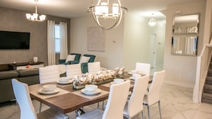 Een eethoek met een houten tafel gedekt voor vier personen, omringd door witte stoelen. Achterin staan twee groene fauteuils, erboven een kroonluchter en aan de muur een flatscreen televisie.