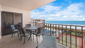 Un balcon avec une table et des chaises, donnant sur une plage et un court de tennis.