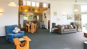 A spacious living area with a blue sofa, a grey sofa, a wooden coffee table, and a dining table with chairs. There is a kitchen area in the background with a window and curtains. - Castlepoint - Private family beach home with stunning views capturing the sun. (Castlepoint)