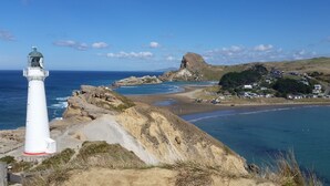 A coastal landscape with a prominent white lighthouse, rocky terrain, and a beach with buildings in the background. - Castlepoint - Private family beach home with stunning views capturing the sun. (Castlepoint)