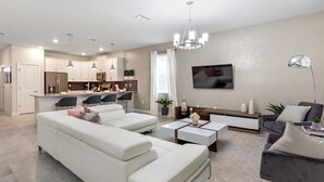 A modern living room with a white sofa, a coffee table, and a grey armchair. There is a kitchen in the background with white cabinets and a dining area.