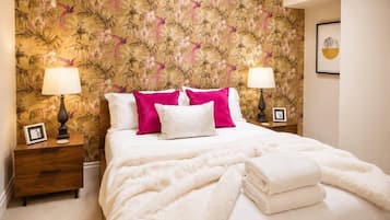 A hotel room with a bed, two bedside tables, a nightstand, a floral wallpaper, and a framed picture on the wall.