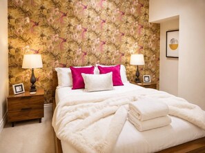 A hotel room with a bed, two bedside tables, a nightstand, a floral wallpaper, and a framed picture on the wall.