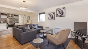 Una sala de estar moderna con un sofá gris, un sillón color beige y una mesa de centro. Se observa una cocina al fondo con gabinetes blancos y electrodomésticos de acero inoxidable.