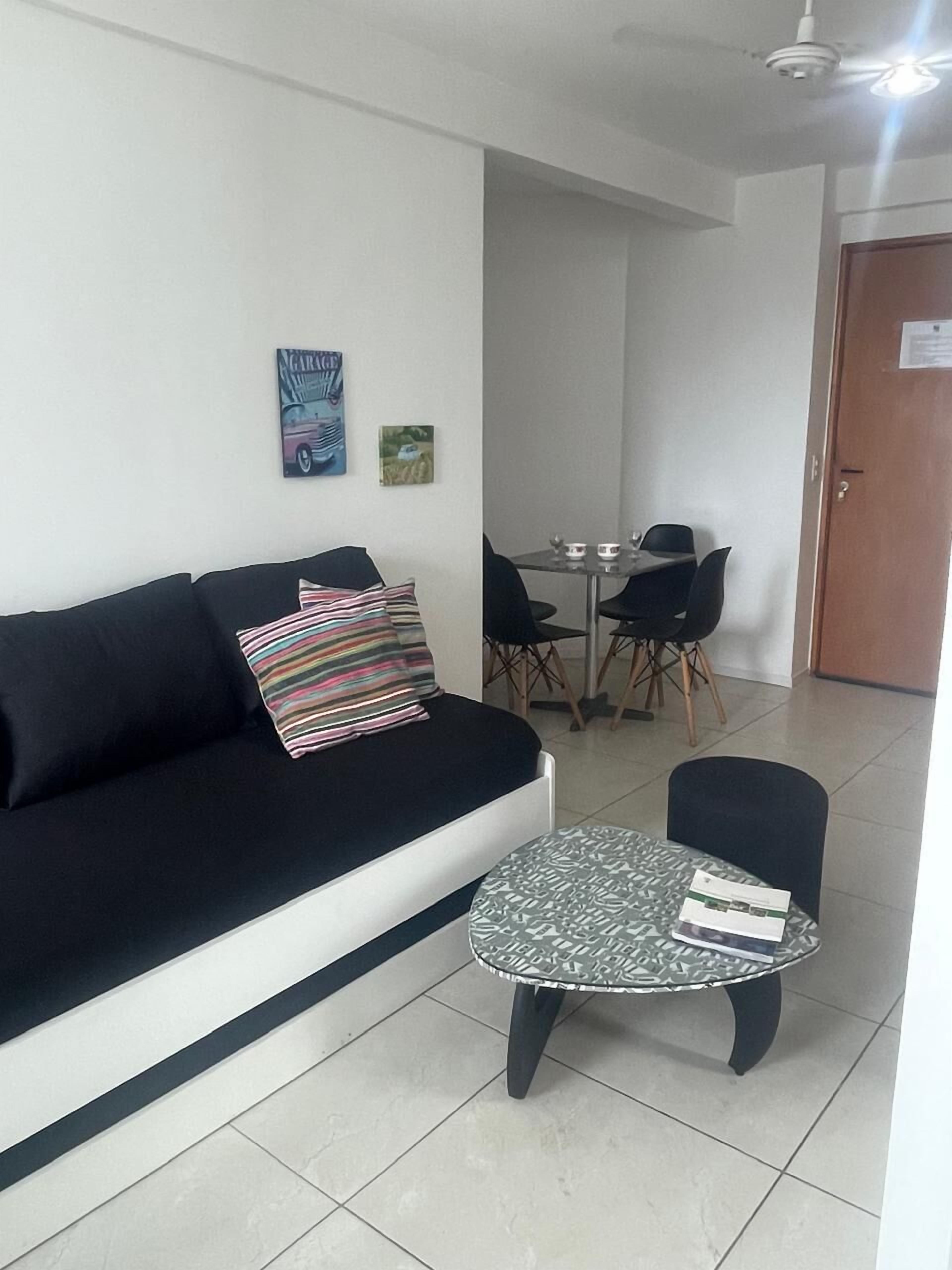 A living room with a black sofa, a striped pillow, a small round table with papers, a dining area with a table and chairs, and a door in the background.
