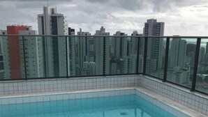 A rooftop swimming pool with tiled edges, surrounded by a glass balustrade, and a cityscape view in the background.