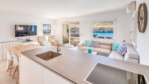 A modern kitchen with a central island, a dining area with white chairs, a sofa with patterned cushions, and a large window with a view of the sea. - Holiday Apartment "Mirador" with Sea Views and Pool (Cala San Vicens)