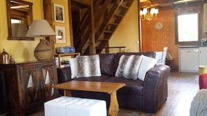 A cozy living room with a leather sofa, a wooden coffee table, and a white ottoman. A wooden cabinet and a lamp are on the left. A staircase leads to an upper level. - Warm Traditional Chalet, Snow / Sun Hautes Alpes, All Comfort (Chabottes)
