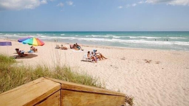 A beach with people relaxing under umbrellas, a wooden structure in the foreground, and the ocean in the background.