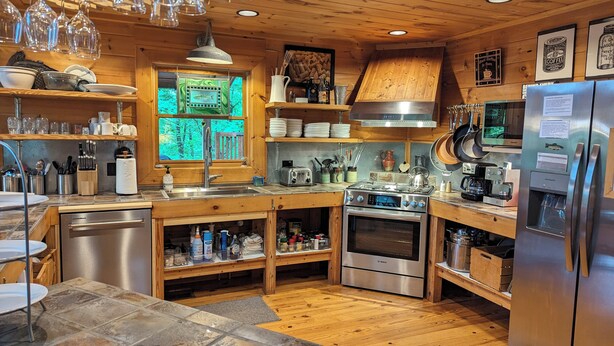 A modern kitchen with wooden cabinets, stainless steel appliances, and a large window with a view of the forest. - Mountain Cabin on Cooper Creek (Suches)