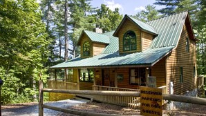 A rustic log cabin with a green metal roof, surrounded by dense forest. - Mountain Cabin on Cooper Creek (Suches)