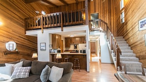 A spacious living area with a high ceiling, wooden walls, a cozy couch, a staircase, and a kitchen area.