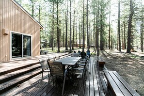 Une terrasse en bois avec une longue table et des bancs, entourée de grands arbres.