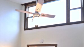 A bedroom with a bed, a wooden dresser, a desk with a lamp, a ceiling fan, and a window.