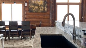 L’intérieur d’une cabane en rondins comprend un coin cuisine avec un évier encastré dans un plan de travail en granit, une table à manger en bois avec des chaises, et un tableau accroché au mur.
