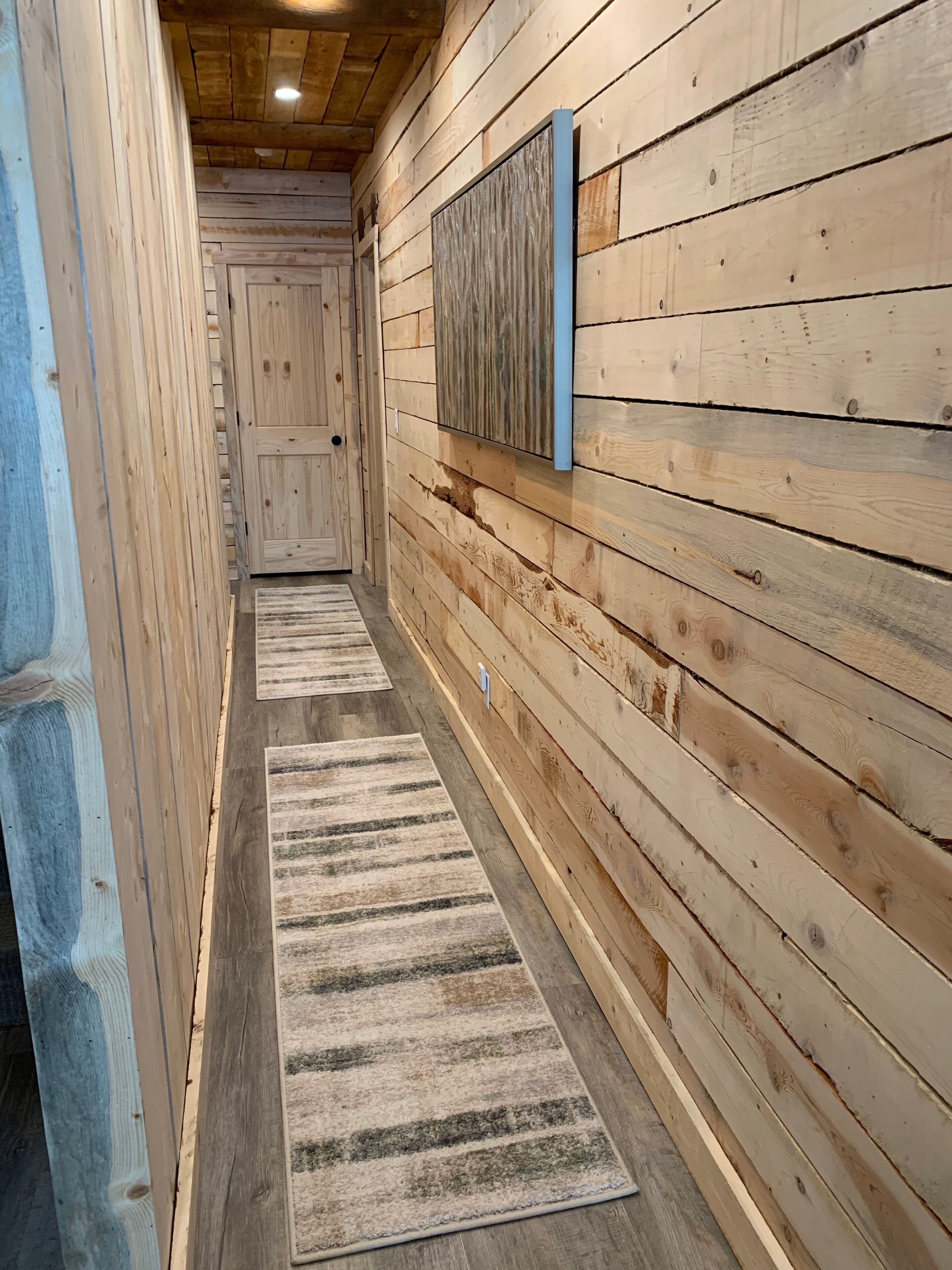 Un pasillo de madera con una puerta al final, un aplique en la pared y dos alfombras en el suelo.