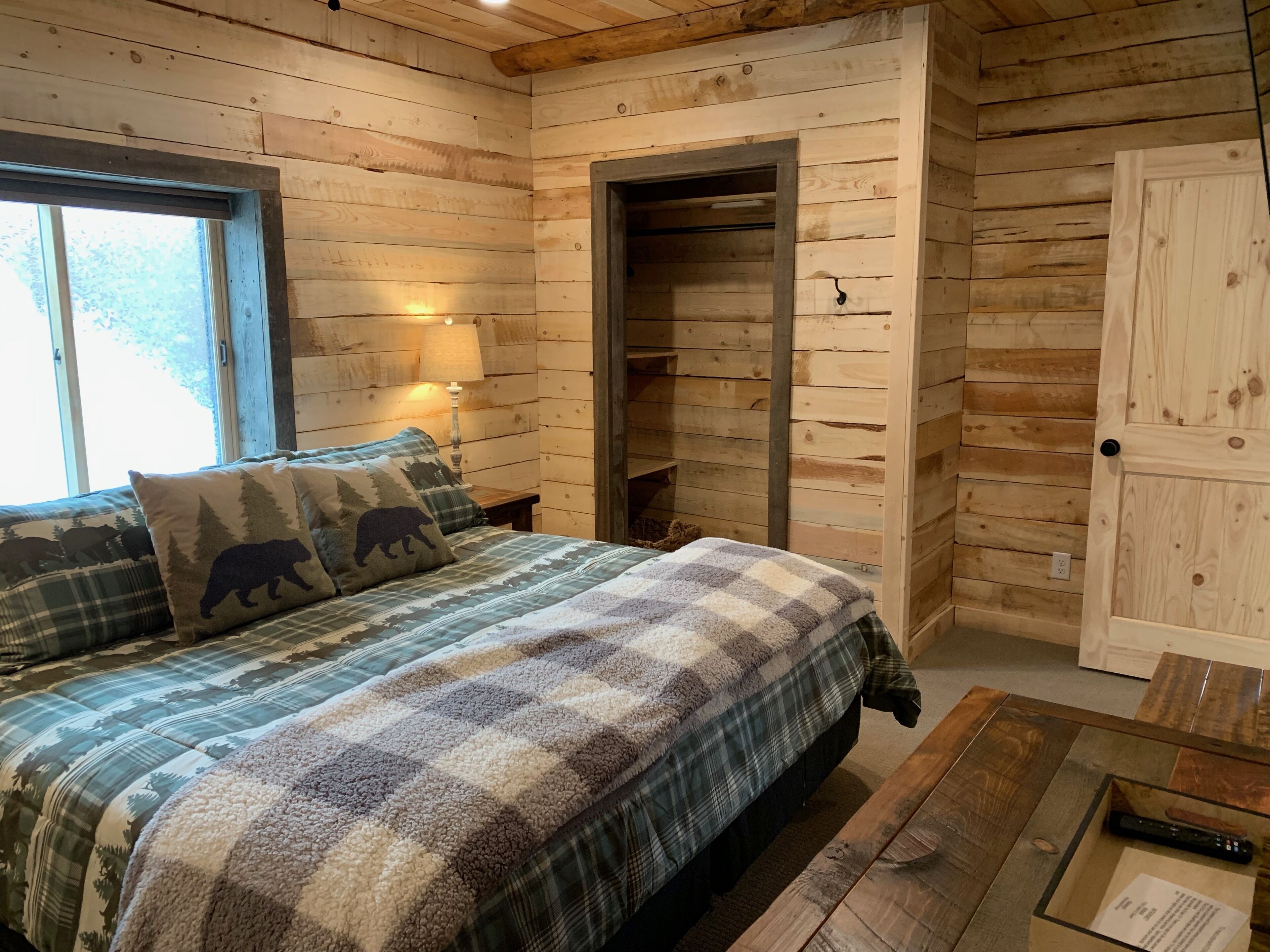 Ein Schlafzimmer in einer Holzhütte mit einem Bett, einem Nachttisch, einer Lampe, einer Holztür und einem Fenster.