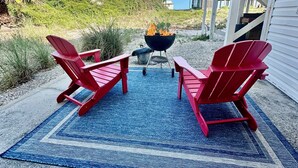 Een terras aan het strand met twee rode Adirondack-stoelen, een zwarte barbecue en een blauw patroon tapijt op een betonnen vloer.