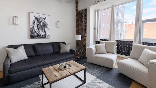 A modern living room with a black sofa, a beige sofa, and a wooden coffee table. There is a large window with curtains, a brick wall, and a mounted artwork of a horse.