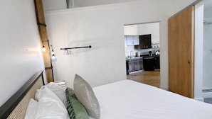 A modern bedroom with a large bed, a wooden headboard, and a view into a kitchen area.