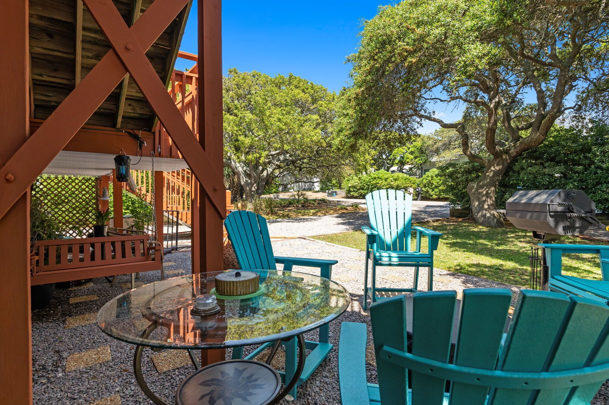 A covered patio with a glass table, green Adirondack chairs, and a barbecue grill.