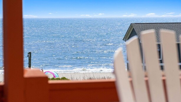 Vista sulla spiaggia con una sedia di legno in primo piano, un edificio con tetto spiovente e cielo sereno.