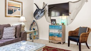 Un soggiorno con un divano, un tavolino da caffè, un televisore a parete, un pesce decorativo, una poltrona in vimini e un mobile con una scena marina.
