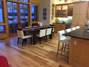 A modern kitchen with wooden cabinets, a dining area with a wooden table and white chairs, and large windows with a view of the outside.