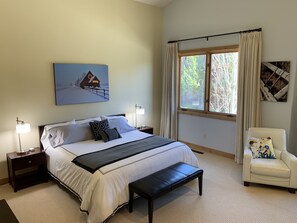 A bedroom with a large bed, a bench, a chair, a nightstand, a lamp, and a window with curtains.