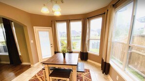 A dining area with a wooden table and bench, large windows with curtains, and a view of a residential area.