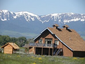 A wooden cabin with a balcony, surrounded by a grassy field and mountains in the background.