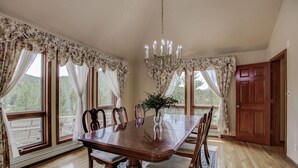 Une salle à manger avec une table en bois, des chaises, un lustre et de grandes fenêtres ornées de rideaux à motifs floraux.