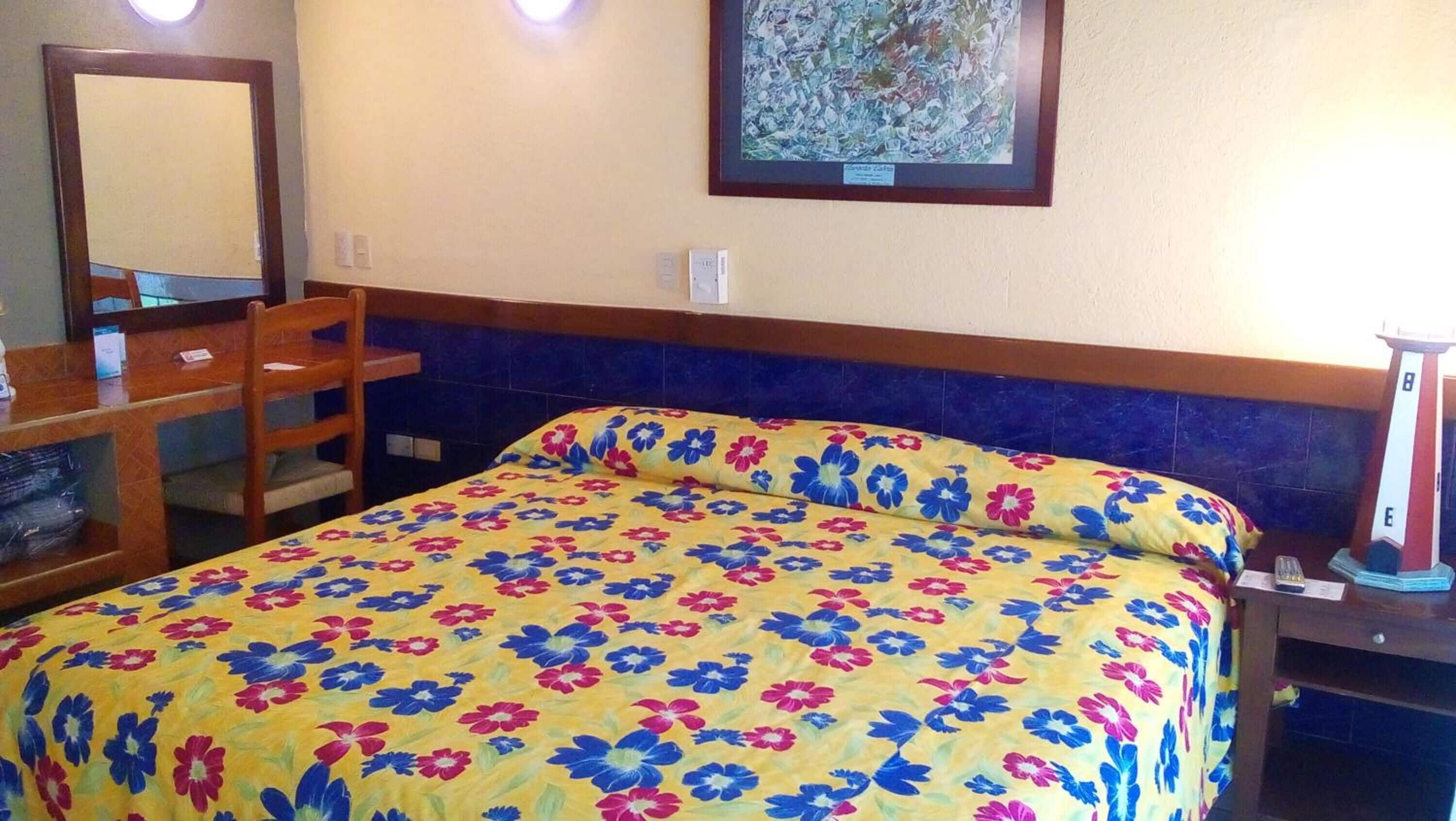 A hotel room with a bed, a desk, a chair, a mirror, and a nightstand.