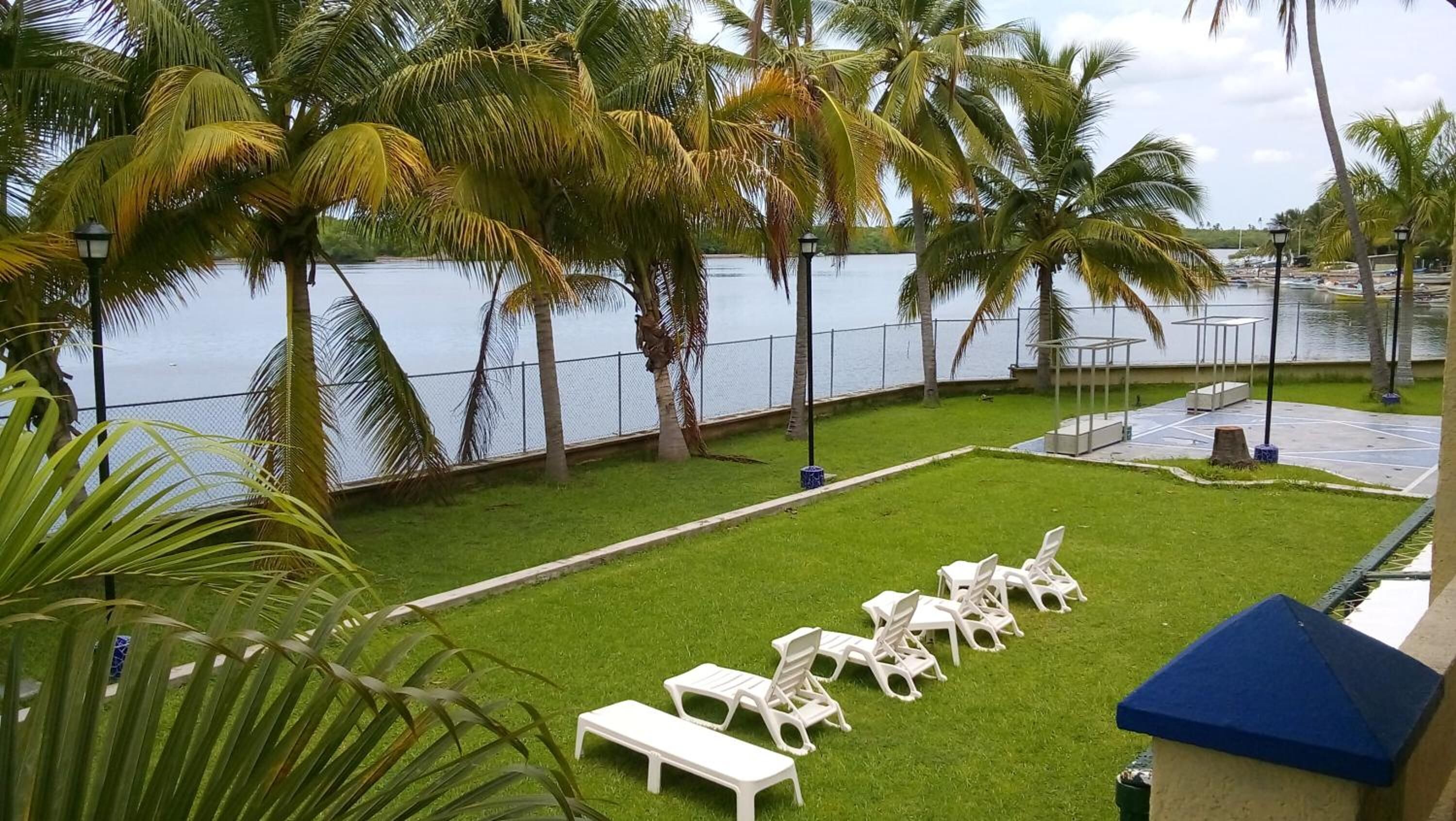 Una zona verde con poltrone bianche e un tavolino a bordo piscina, circondata da palme e vista sull'acqua.