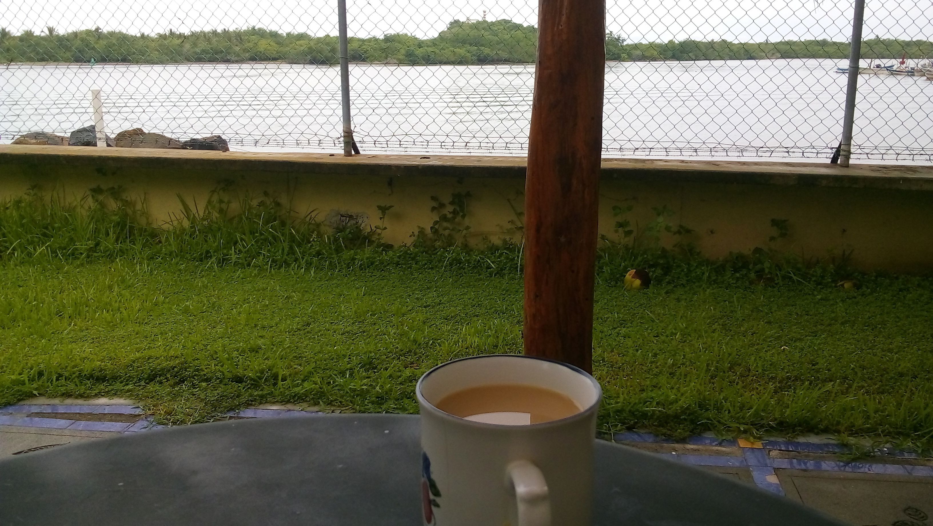 A table with a cup of coffee, a chain-link fence, and a view of a body of water with boats.