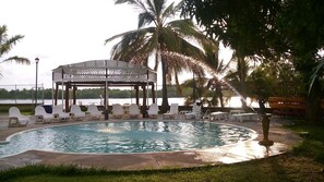 A circular pool with lounge chairs and a gazebo, surrounded by palm trees and a view of a body of water.