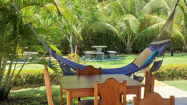 A hammock chair under a wooden pergola, a wooden table, and chairs in a garden with palm trees and a fountain. - Beautiful Condominium - Wifi access throughout and 300 Yards from the Beach! (Samara)