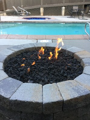 A fire pit with flames, surrounded by stone blocks, near a swimming pool with a wooden fence and outdoor furniture in the background. - Highest Rated, Lowest Rates in Snow Blaze: 2/2, A/C, Pool, Hot Tub, Gym, Firepit (Park City)