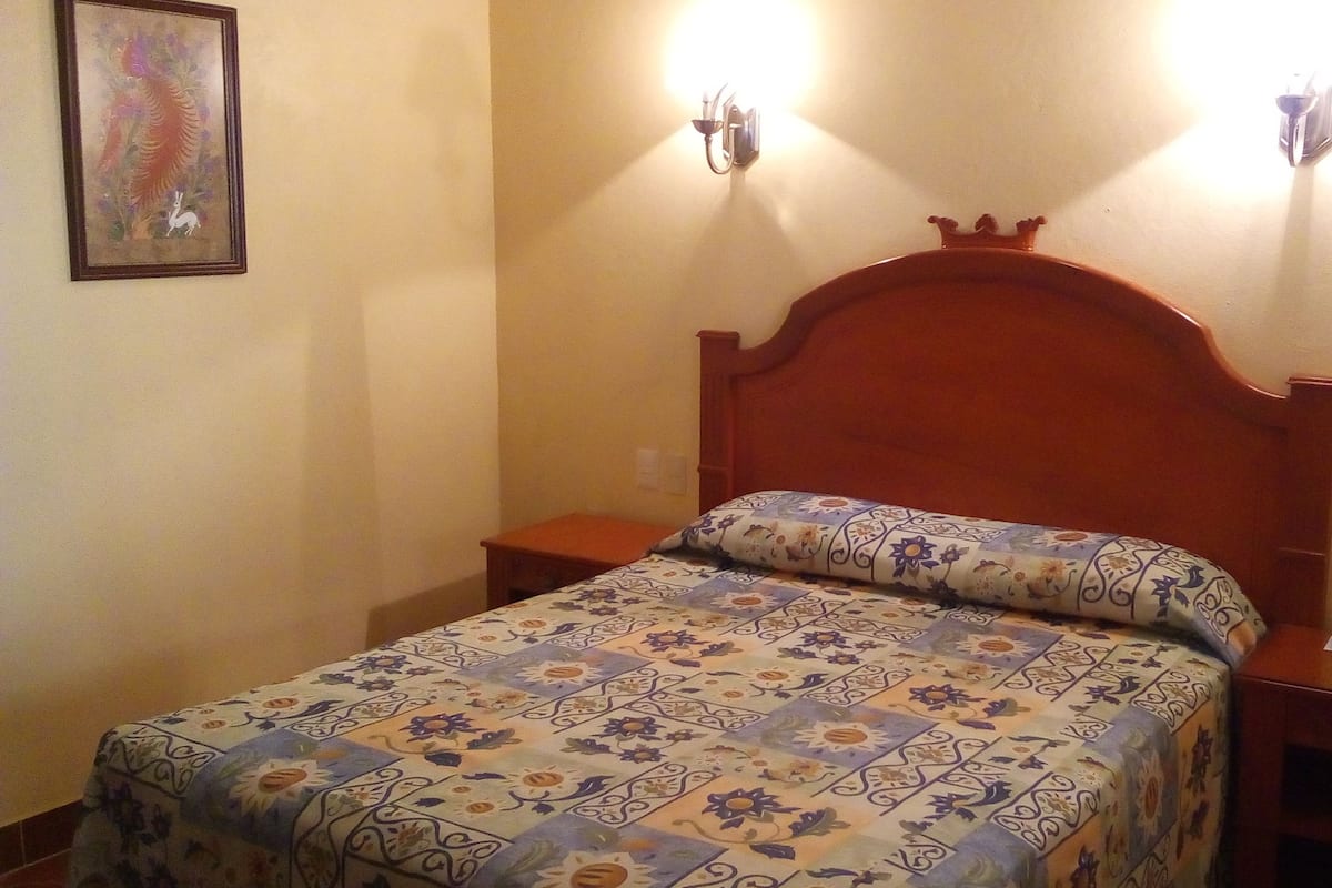 Un dormitorio con cabecera de madera, colcha estampada con motivos florales, dos lámparas de noche y una mesita de noche con control remoto.