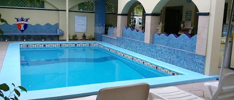 Une piscine dont les bords sont recouverts de carreaux et qui est agrémentée d’un mur décoratif. On y trouve des chaises longues et un petit coin salon avec une fresque murale.