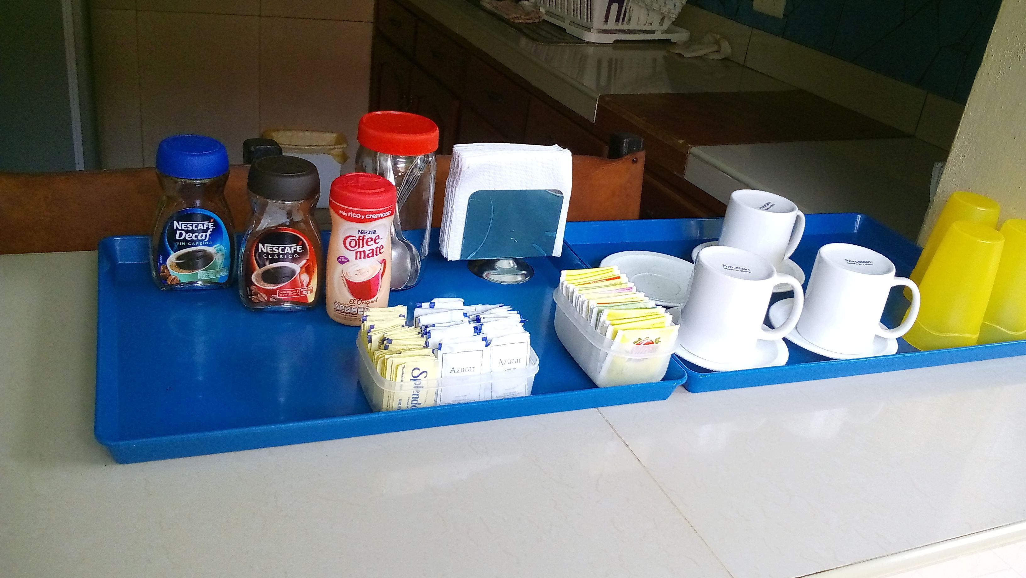 Una barra de desayuno con diversas opciones de café y té, incluyendo Nescafé Decaf, Nescafé Classic y Nescafé Coffee-Mate. También hay sobres de azúcar, una cafetera y tazas y platillos blancos.