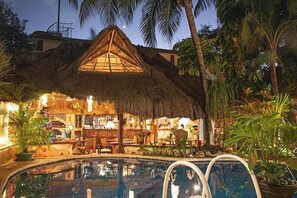 Un espacio al aire libre con techo de paja, una piscina, sillones de descanso y plantas tropicales.