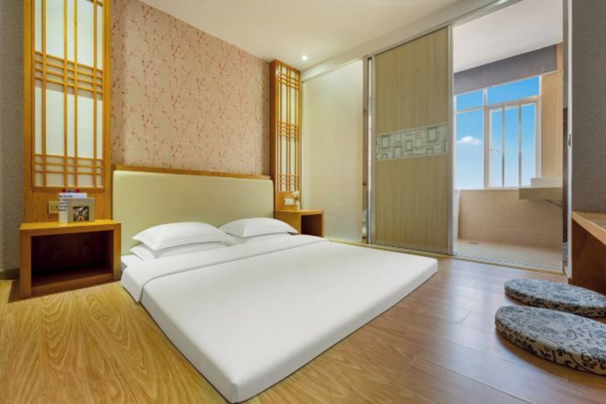A modern bedroom with a large bed, wooden headboard, and a sliding glass door leading to a balcony.