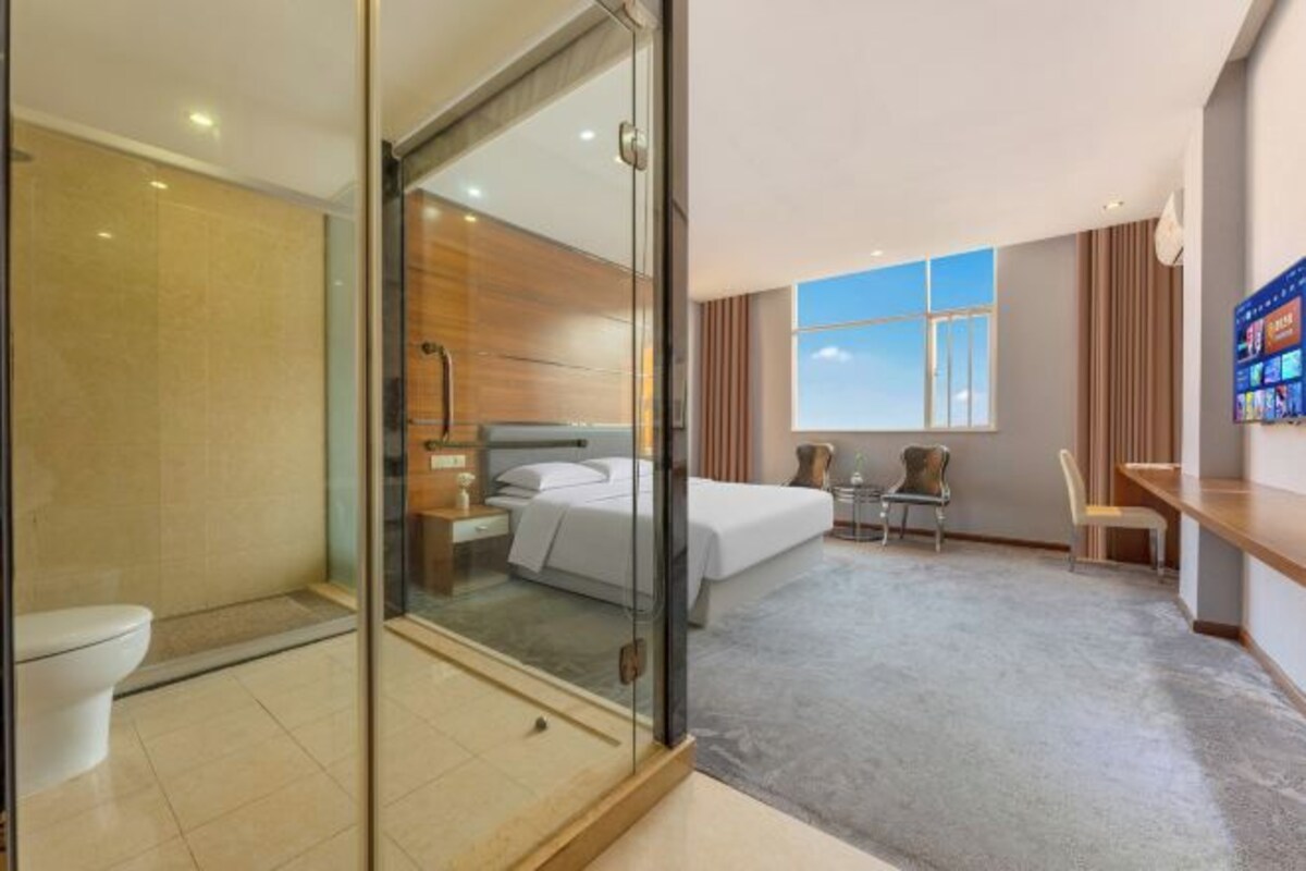 A hotel room with a glass partition separating the bathroom from the bedroom, a bed, a desk, and a chair.